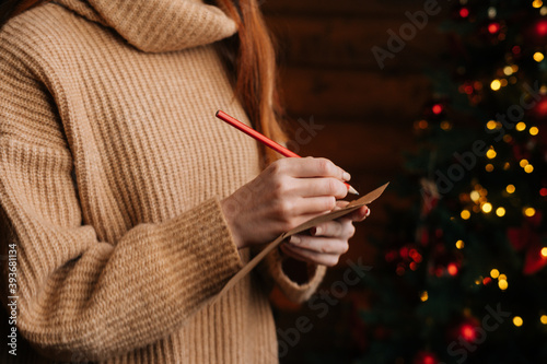 Close-up hands of unrecognizable young woman writing Christmas letter to Santa Claus at cozy dark living room. Xmas tree baubles and tinsel lights blinking at New Year Eve on blur bokeh background.