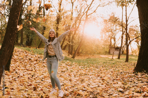 Full length of a cute young teenage caucasian girl with a hat in jeans throwing leaves in the park. Sunny autumn day in the woods