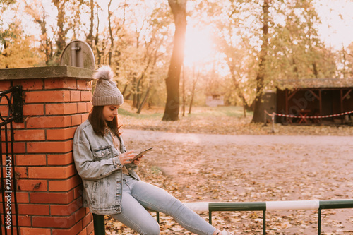 A young modern caucasian girl with a hat dressed in jeans sits in the park and uses the phone. Hello November