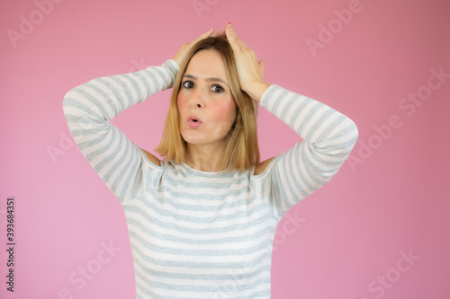 Portrait of young woman with blond hair touching her head and expressing worry isolated over pink background