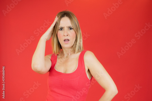 Portrait of young woman with blond hair touching her head and expressing worry isolated over red background
