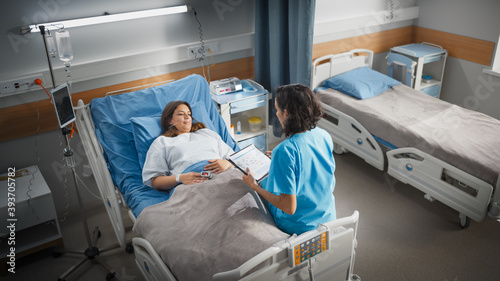 Hospital Ward  Friendly Hispanic Head Nurse Fills Medical History Form  Talks to Female Patient Recovering in Bed. Professional Nurse Helps Happy Woman Get Better after Surgery. High Angle Shot