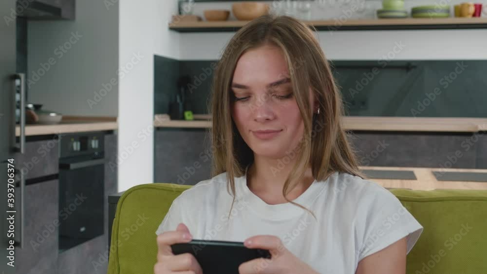 Smiling young woman playing cellphone app game at living room. Young caucasian female enjoying smartphone video games while sitting on sofa at living room. Smiling young woman playing cellphone app game at living room. Young caucasian female enjoying smartphone video games while sitting on sofa at living room.