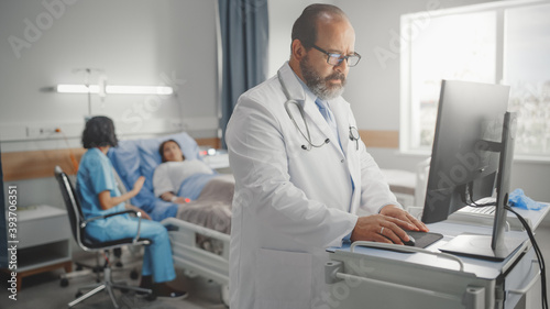 Hospital Ward: Professional Experienced Latin Doctor / Surgeon Uses Medical Personal Computer. In the Background Head Nurse Sitting with a Patient Recovering After Surgery in Bed 