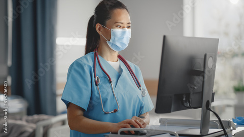 Hospital Ward: Professional Experienced Chinese Head Nurse / Doctor Wearing Face Mask Uses Medical Touch Screen Computer, Checking Patient's Medical Data. In the Background Patient Recovering on Bed. photo