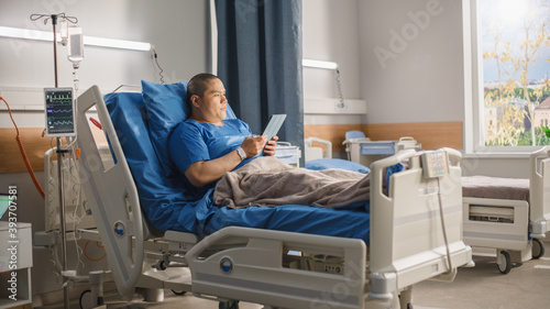 Medical Hospital Ward: Portrait of Handsome Chinese Man Resting in Bed, Working on Digital Tablet Computer, Communicating with Family and Friends, e-business. Businessman Recovering after Surgery
