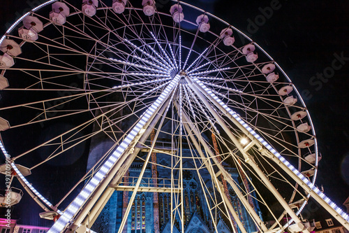 A ferris wheel sparkling with neon signs, and a cathedral decorated with Christmas decorations visible between it. Mulhouse, france