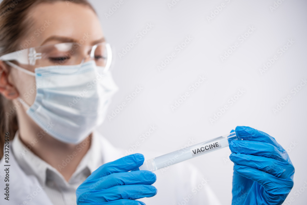 Close-up view of female microbiologist in blue protective gloves, glasses and face mask look at biological tube with covid-19 vaccine on white background. Research and and development concept. Close-up view of female microbiologist in blue protective gloves, glasses and face mask look at biological tube with covid-19 vaccine on white background. Research and and development concept.