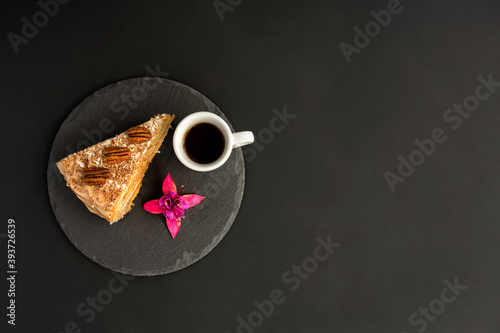 A typical Portuguese cake made of cookies called Bolo de Bolasa with cream, pecans, black and white chocolate on a black slate with espresso and a flower on a black background. Flatlay. Copy space