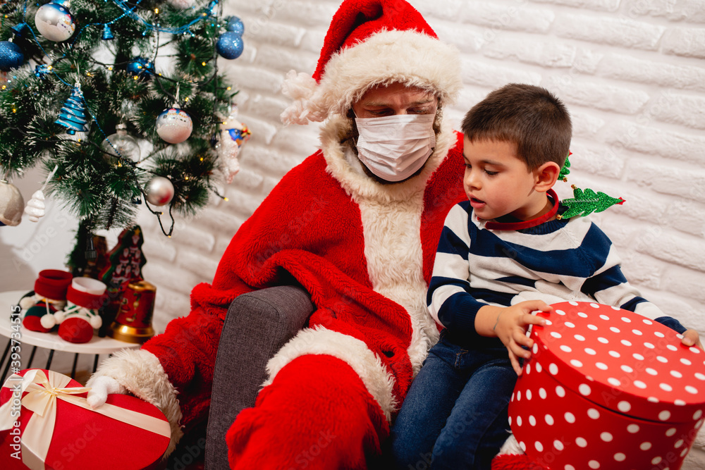 A 5 year old boy sitting in Santa's lap with a face mask and watching a xmas gift next to a Christmas tree. New Year and Christmas pandemic COVID -19 coronavirus A 5 year old boy sitting in Santa's lap with a face mask and watching a xmas gift next to a Christmas tree. New Year and Christmas pandemic COVID -19 coronavirus