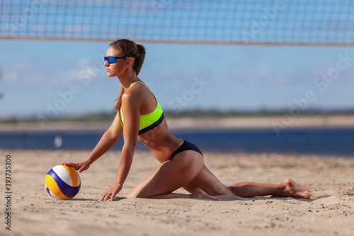Back view of sexy athletic girl in a swimsuit and sunglasses touches a ball while sitting on the sand next to a beach volleyball net © satyrenko