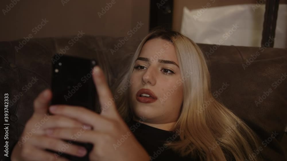 Young blond beautiful woman stretching on the sofa is playing game with smartphone and feeling sad Young blond beautiful woman stretching on the sofa is playing game with smartphone and feeling sad