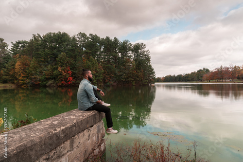 a young man sits on ledge looking out over a lake on a cloudy fall afternoon