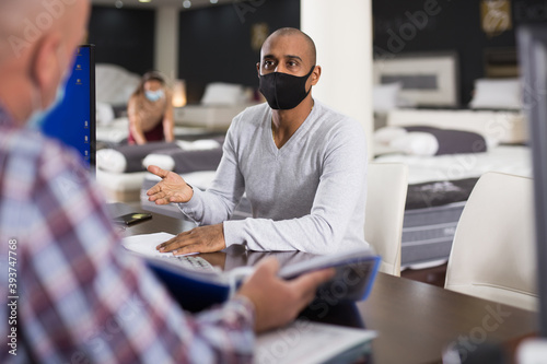 Latin american man in protective mask visiting furniture store, talking to salesman. Concept of prevention and social distancing in coronavirus pandemic