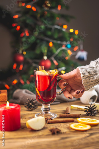 A woman's hand in a warm sweater is holding a cup of aromatic hot mulled wine against the background of a Christmas tree with lights. Concept of a festive atmosphere