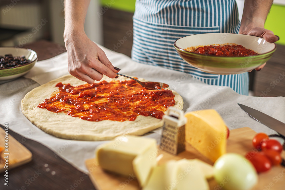 Female chef in an apron is putting tomato sauce on a raw pizza. Cooking delicious pizza at home in the kitchen.