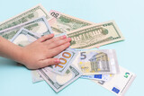 Top view of a child's hand on the background of money dollars and euros on a pastel blue background, top view