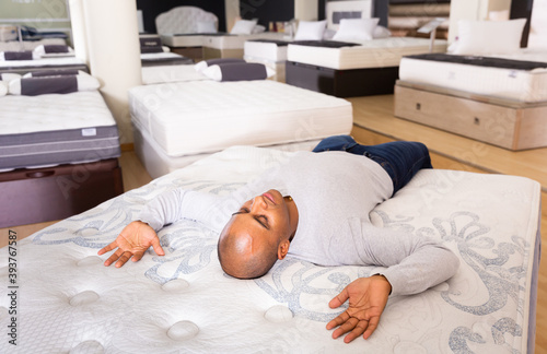Smiling adult hispanic man lying on bed in furniture store, enjoying convenience of modern orthopedic mattress .