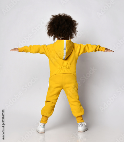 Curly dark-skinned kid girl in warm yellow sports jumpsuit and sneakers stands back to camera holding arms up spread wide over white wall background. Trendy children fashion, stylish outfit