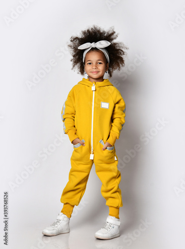Happy smiling dark-skinned kid girl in warm yellow jumpsuit and sneakers stands holding hands in her pockets