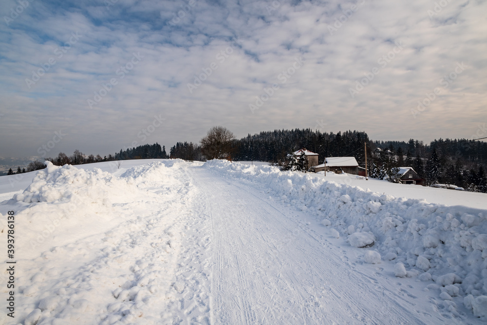 winter scenery with snow covered road, few houses, smaller hills and blue sky with clouda above Bukovec village in Czech republic