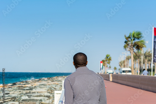 An African man watching the seascape at the crescent road and the breakwater and stones and background of Persian gulf in the Palm Jumeirah island in Dubai of the United Arab Emirates.