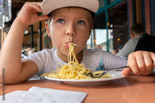 a boy in a cap 6 years old is sitting in a cafe and eating spaghetti
