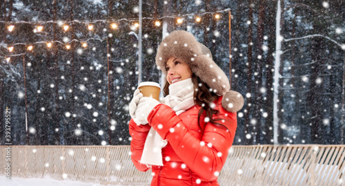 christmas, hot drinks and holidays concept - happy woman with disposable paper coffee cup in winter over outdoor ice skating rink on background