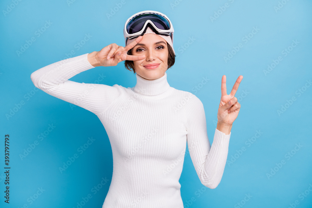 Photo of charming young woman show v-sign good mood winter season skier isolated on blue color background Photo of charming young woman show v-sign good mood winter season skier isolated on blue color background