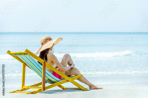 Beautiful Asian woman in bikini and sun hat sitting on beach chair and applying sunscreen for sunbathing on island beach. Pretty girl relax and enjoy with outdoor lifestyle and summer holiday vacation