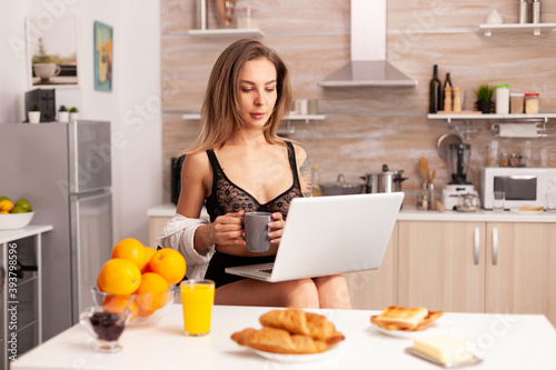 Attractive blonde woman in sexy lingerie holding coffee while using laptop in home kitchen. Beautiful lady with tattos during breakfast in underwear.