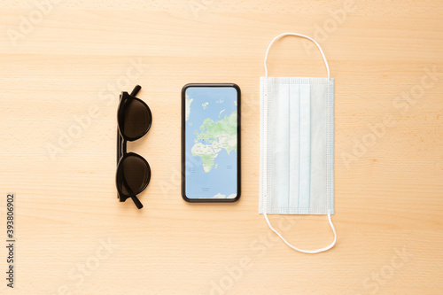 Flat lay, travel concept during the Covid-19 outbreak, a face mask, sunglasses and a smartphone with a World Map are arranged on a wooden table. 