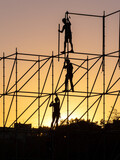 Builders are dismantling the tubular structure. Against the backdrop of the setting sky, only silhouettes are visible. Workers hold tools and pipes.