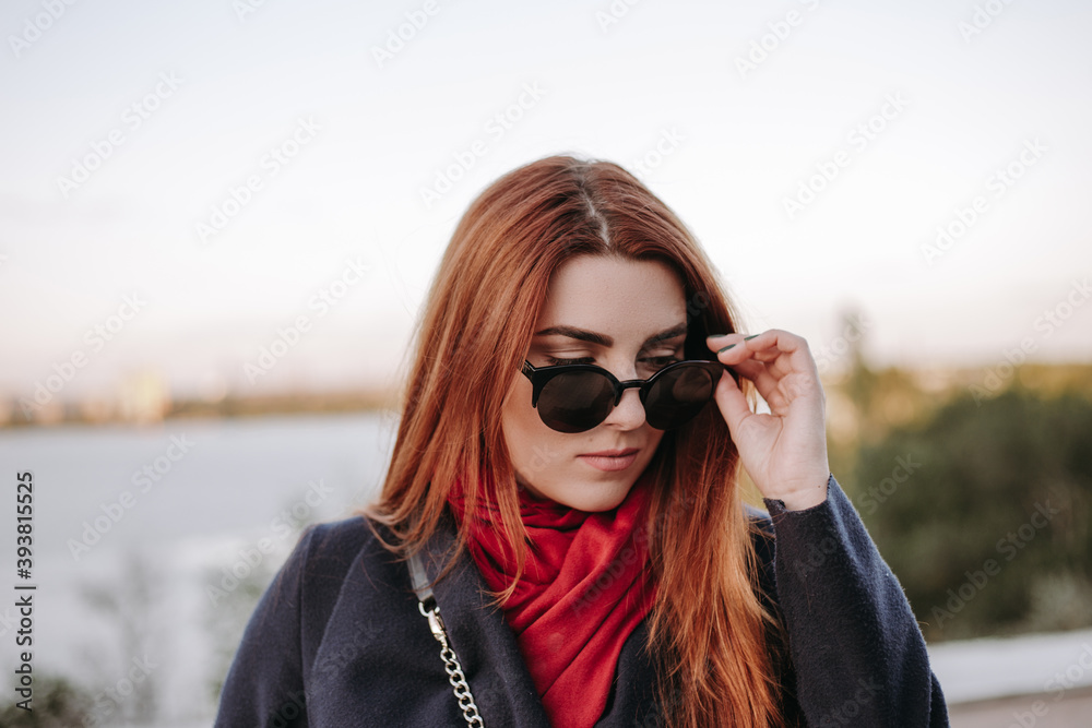Pretty woman with green eyes and wide eyebrows and red hair posing on the city street and smiling. Model dressed in warm clothes. Red scarf black coat and sunglasses. Romantic pretty female model Pretty woman with green eyes and wide eyebrows and red hair posing on the city street and smiling. Model dressed in warm clothes. Red scarf black coat and sunglasses. Romantic pretty female model