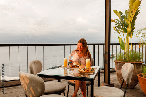 Young woman in a fine dining restaurant eat a hamburger with salad and french fries, enioy sunset by the sea photo