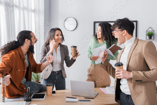 Excited hispanic co-workers looking at female colleague talking on mobile phone near workplace photo