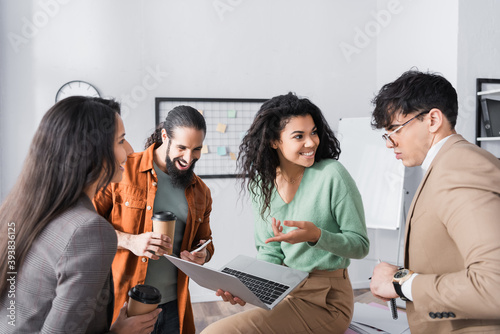 Smiling hispanic woman pointing with finger at laptop, while looking at co-worker during break at workplace