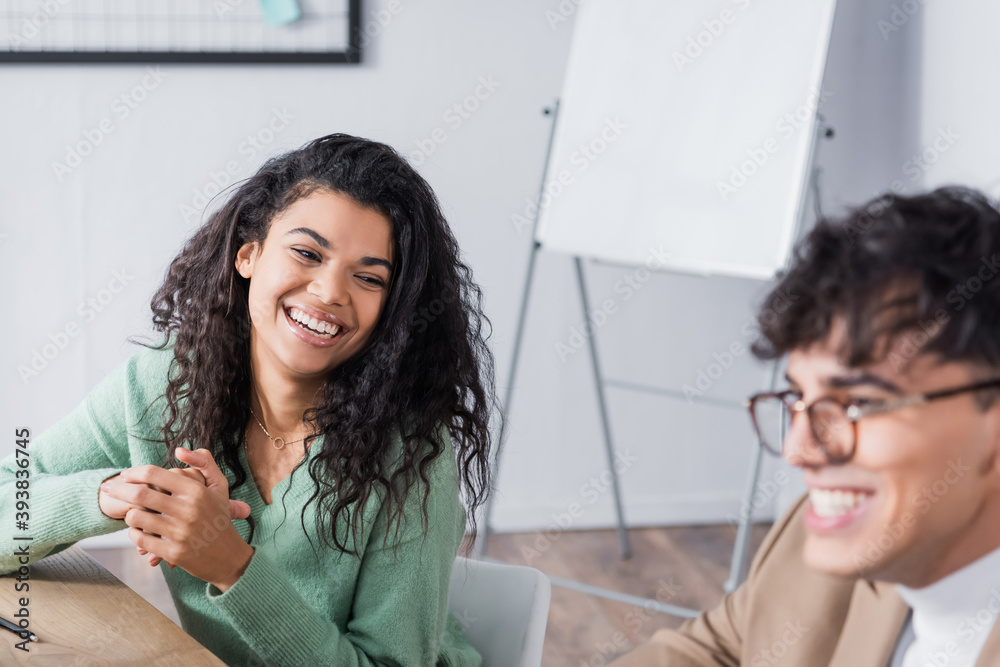 Fototapeta premium Curly hispanic woman laughing, while looking at co-worker sitting at desk in office on blurred foreground
