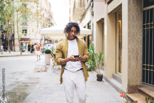 Handsome young male with curly hair using mobile phone on urban settings sending text messages and mails, positive 20s hipster guy browsing web page on smartphone searching destinations and location