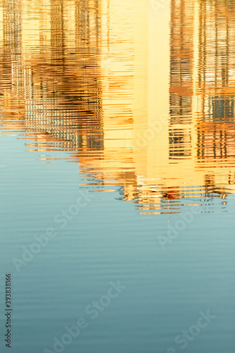 Abstract reflection in the water. Sunny day. Distorted view with ripples. Urban shot. Buildings enlightened by yellow sunset light and blue sky in reflection.