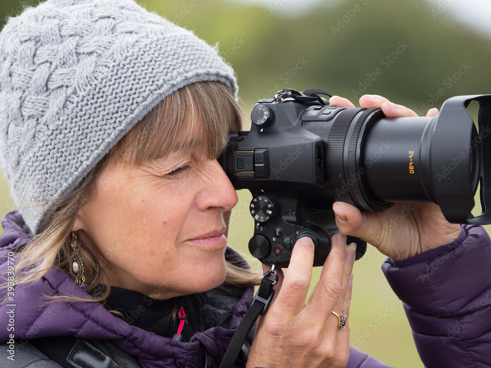 Obraz premium A lady photographer holds a camera in portrait mode to her eye and closes the other eye as she takes a shot.Her finger is on shutter release button.Aperture priority has been selected. Zoom lens