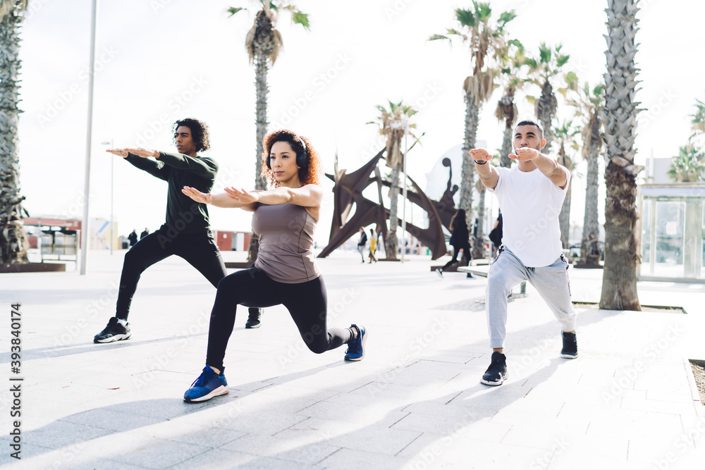 Obraz premium Group of muscular athletes doing squatting exercise during morning workout at urban setting with palm trees, slim female aerobic trainer in bluetooth headphones working with determined male group