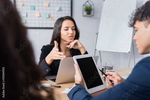 Hispanic businesswoman pointing with hand near colleague with digital tablet on blurred foreground