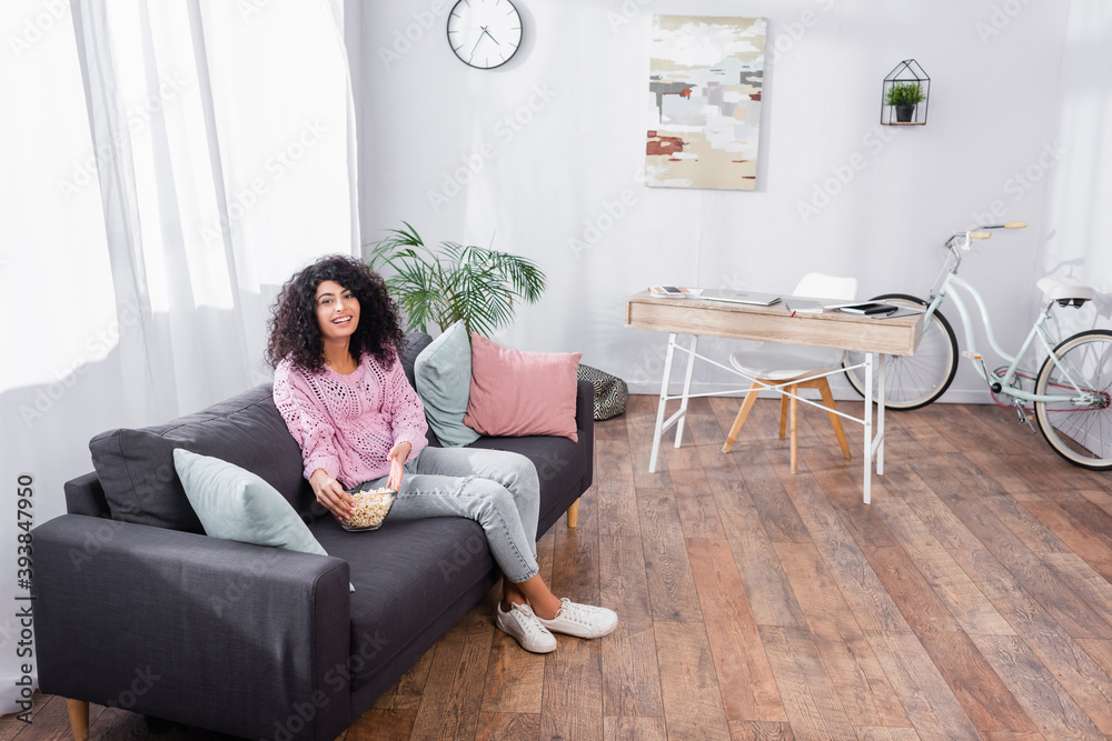cheerful woman sitting on couch with popcorn in bowl in modern living room cheerful woman sitting on couch with popcorn in bowl in modern living room