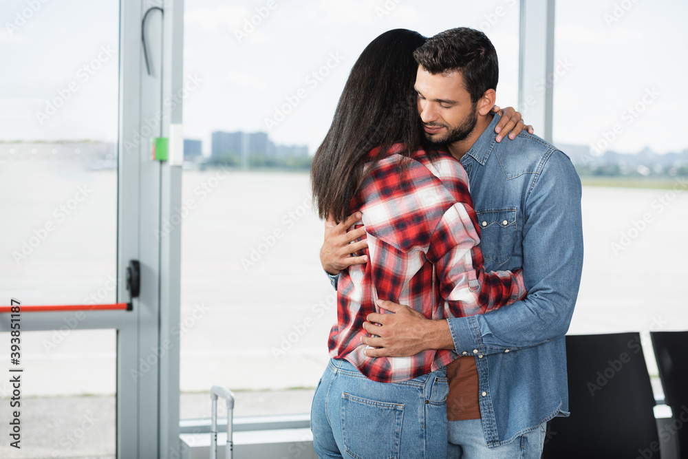 Obraz premium bearded man hugging brunette african american girlfriend in airport