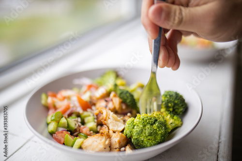 Hand sticking fork on broccoli and chicken salad next to a window
