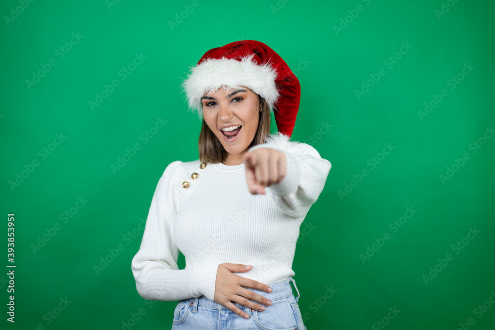 Young beautiful woman wearing a Santa hat and white sweater over green background laughing at you, pointing finger to the camera with hand over body, shame expression Young beautiful woman wearing a Santa hat and white sweater over green background laughing at you, pointing finger to the camera with hand over body, shame expression