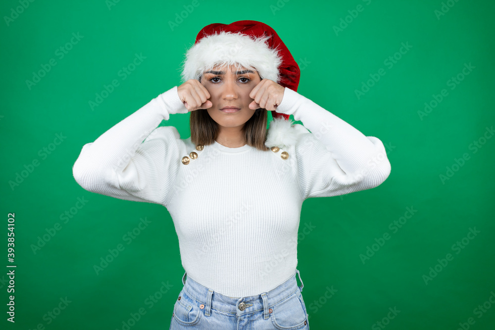 Young beautiful woman wearing a Santa hat and white sweater over green background depressed and worry for distress, crying angry and afraid. Sad expression. Young beautiful woman wearing a Santa hat and white sweater over green background depressed and worry for distress, crying angry and afraid. Sad expression.