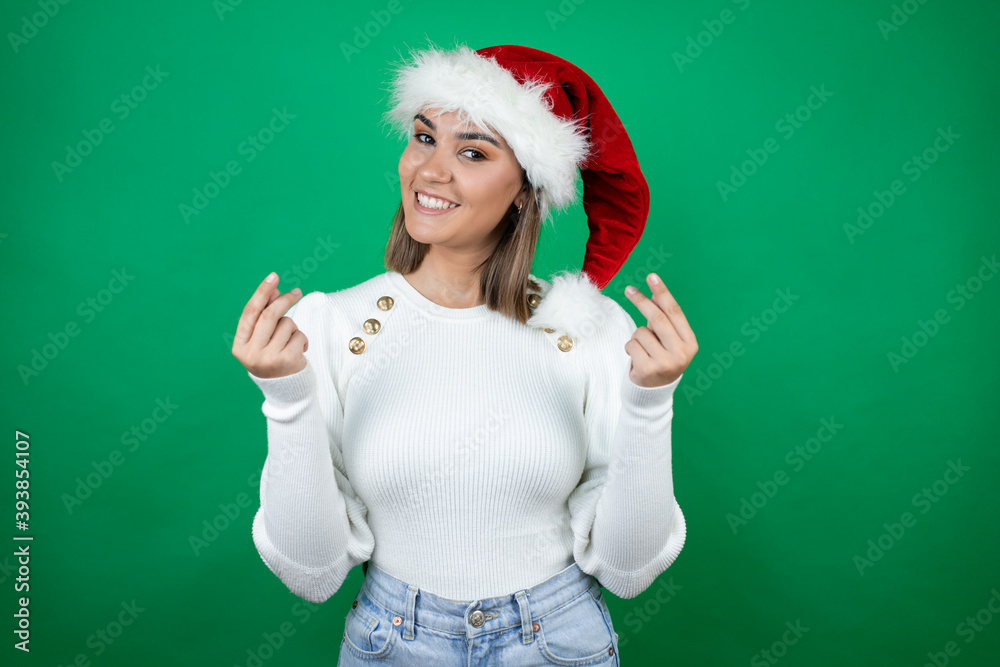 Young beautiful woman wearing a Santa hat and white sweater over green background doing money gesture with hands, asking for salary payment, millionaire business Young beautiful woman wearing a Santa hat and white sweater over green background doing money gesture with hands, asking for salary payment, millionaire business