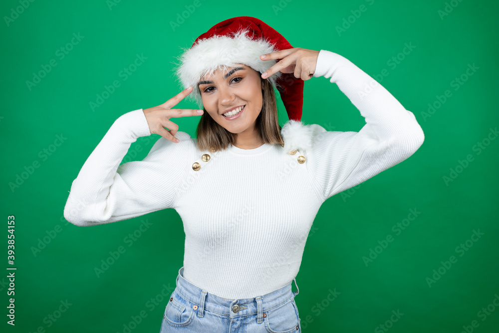 Young beautiful woman wearing a Santa hat and white sweater over green background Doing peace symbol with fingers over face, smiling cheerful showing victory Young beautiful woman wearing a Santa hat and white sweater over green background Doing peace symbol with fingers over face, smiling cheerful showing victory